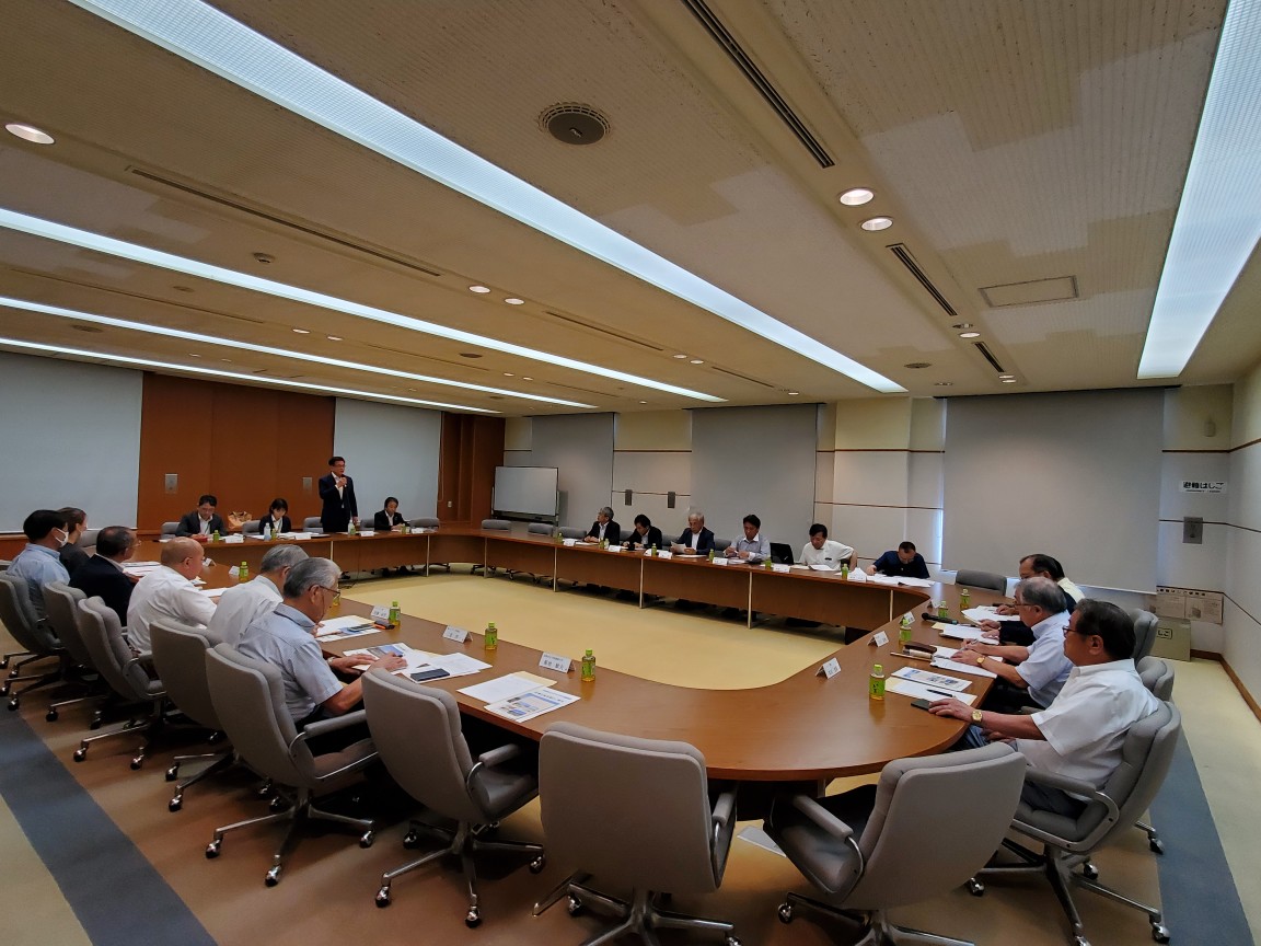 複数の男性がロの字型に並んだ机に座っており、中央に立ったスーツ姿の市長が、資料を指差しながらスピーチをしている様子の写真