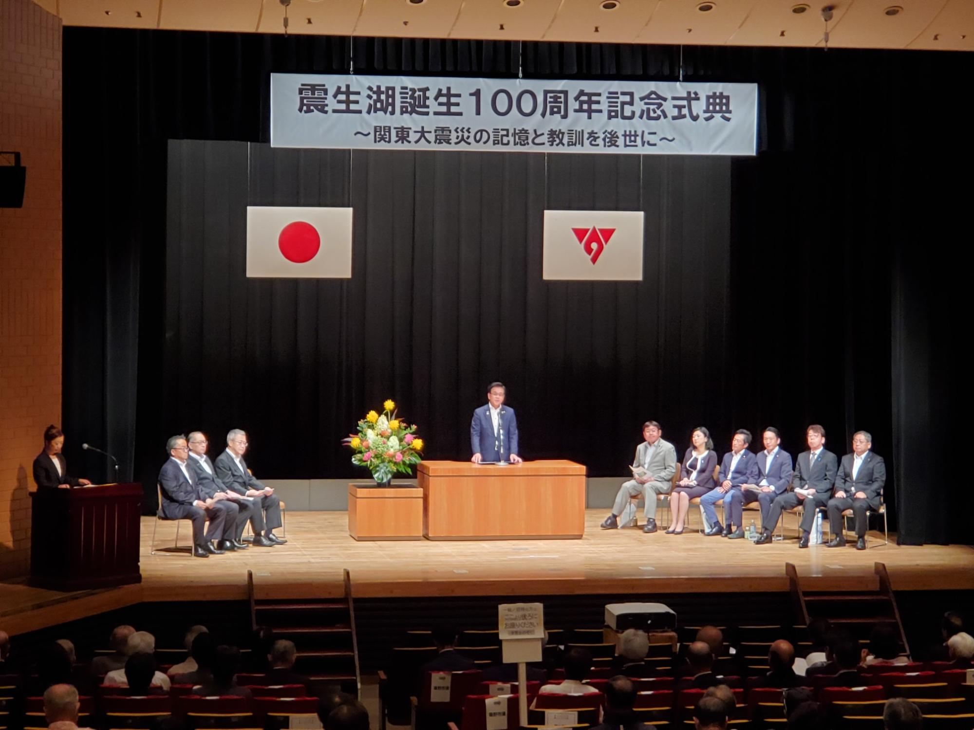 「震生湖誕生100周年記念式典」と書かれた看板が掲げられた舞台で、演説台の前にスーツ姿の市長が立ち話をしている、両脇には数名の関係者や日の丸などの旗が並んでいる記念式典の写真