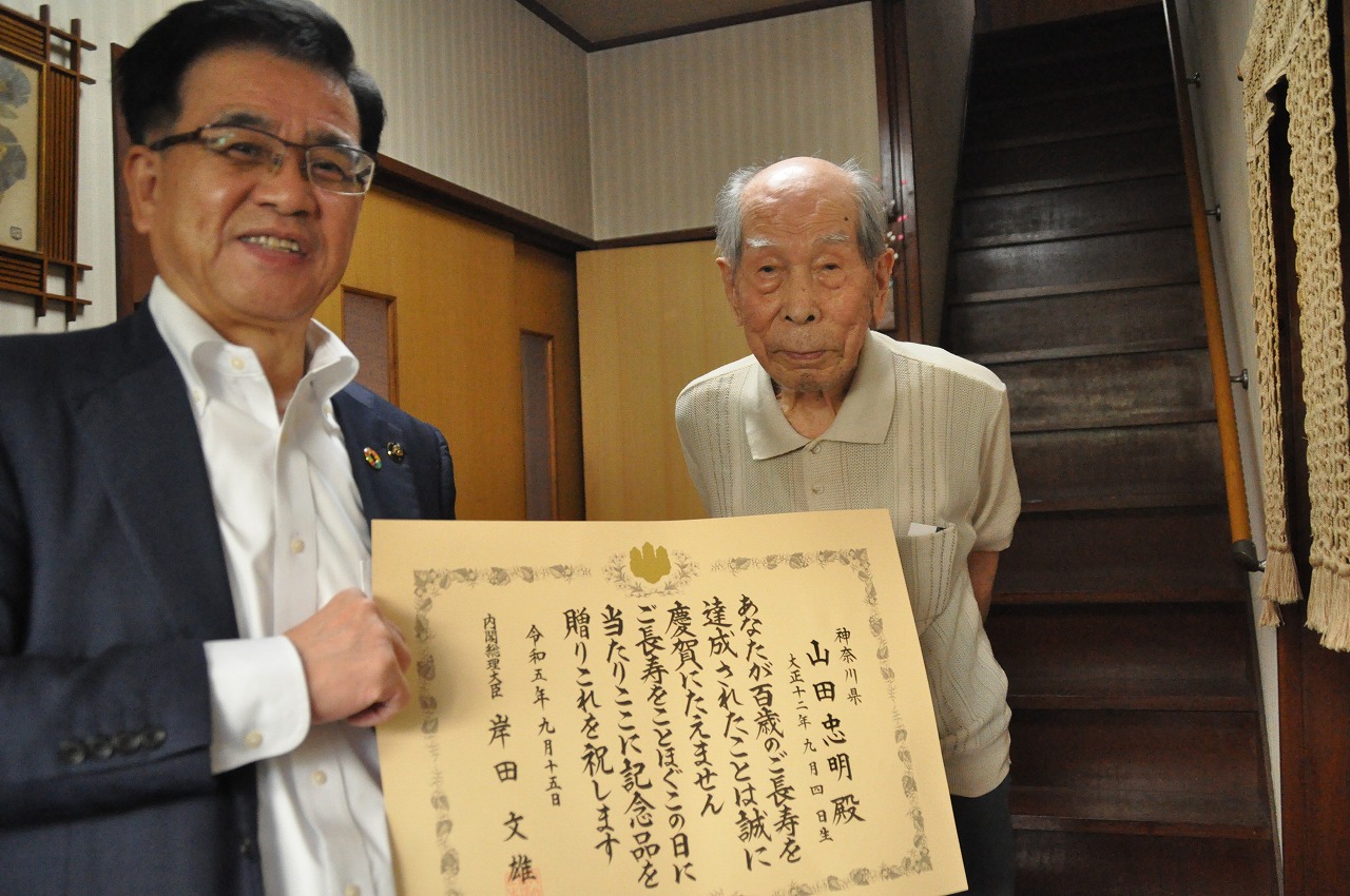 賞状を掲げたスーツ姿の市長と、その横に立つ高齢の男性がカメラに向かって微笑み記念撮影している写真