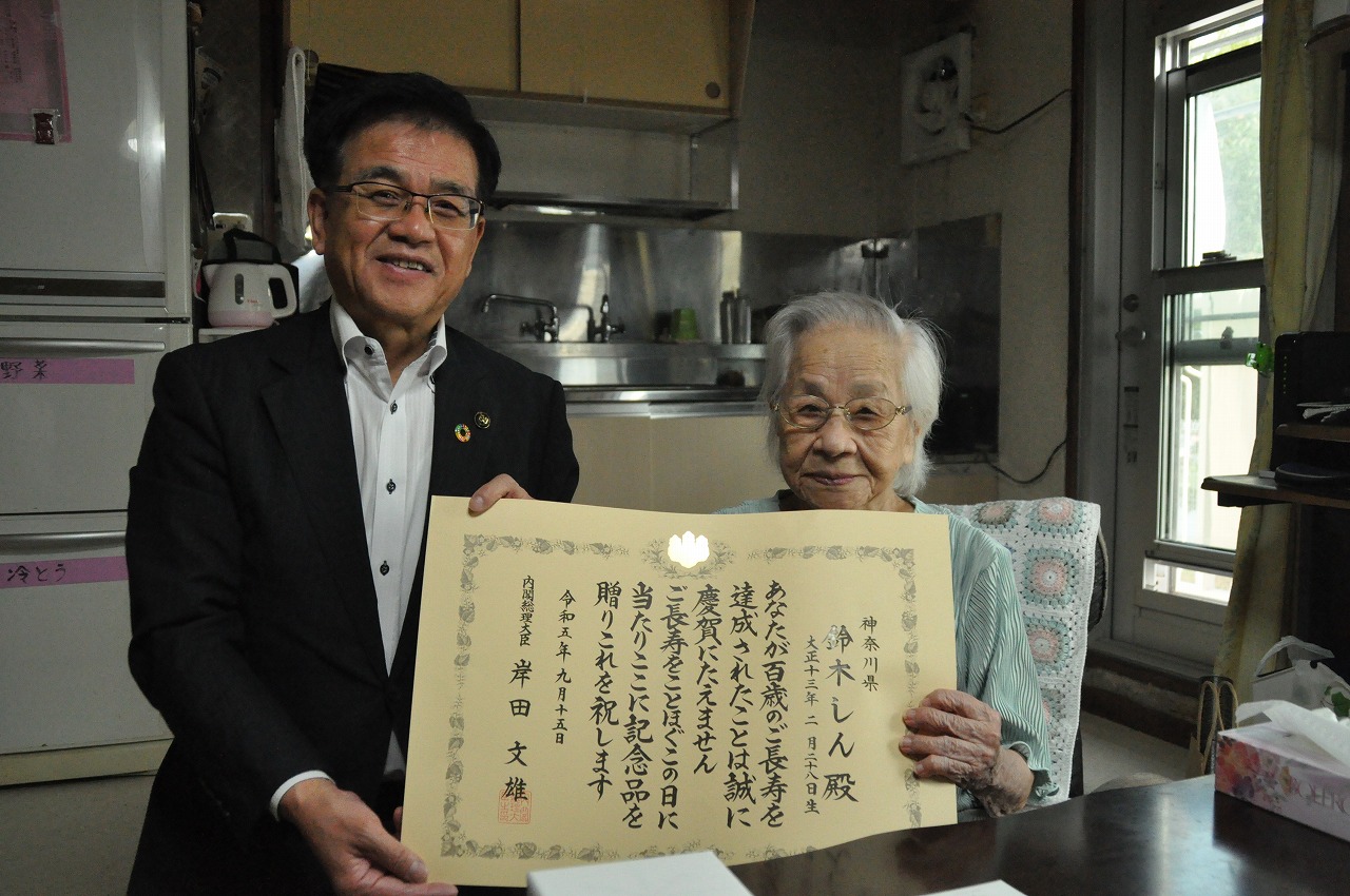 黒いスーツの市長と椅子に座った高齢女性が、部屋の中で賞状を広げて一緒に持ち笑顔で記念撮影している写真