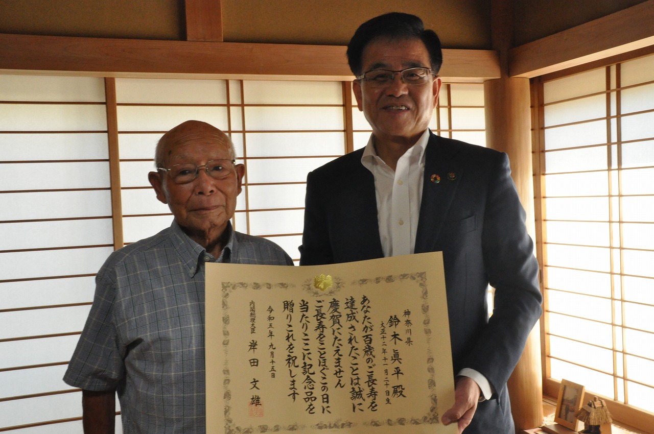 和風の部屋で、スーツを着た市長と高齢の男性が一緒に賞状を持ち記念撮影している写真