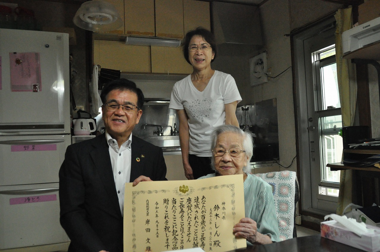 キッチンらしき室内で、椅子に座った高齢女性とスーツ姿の市長が一緒に賞状を持ち、ご家族の女性と一緒に記念撮影している写真