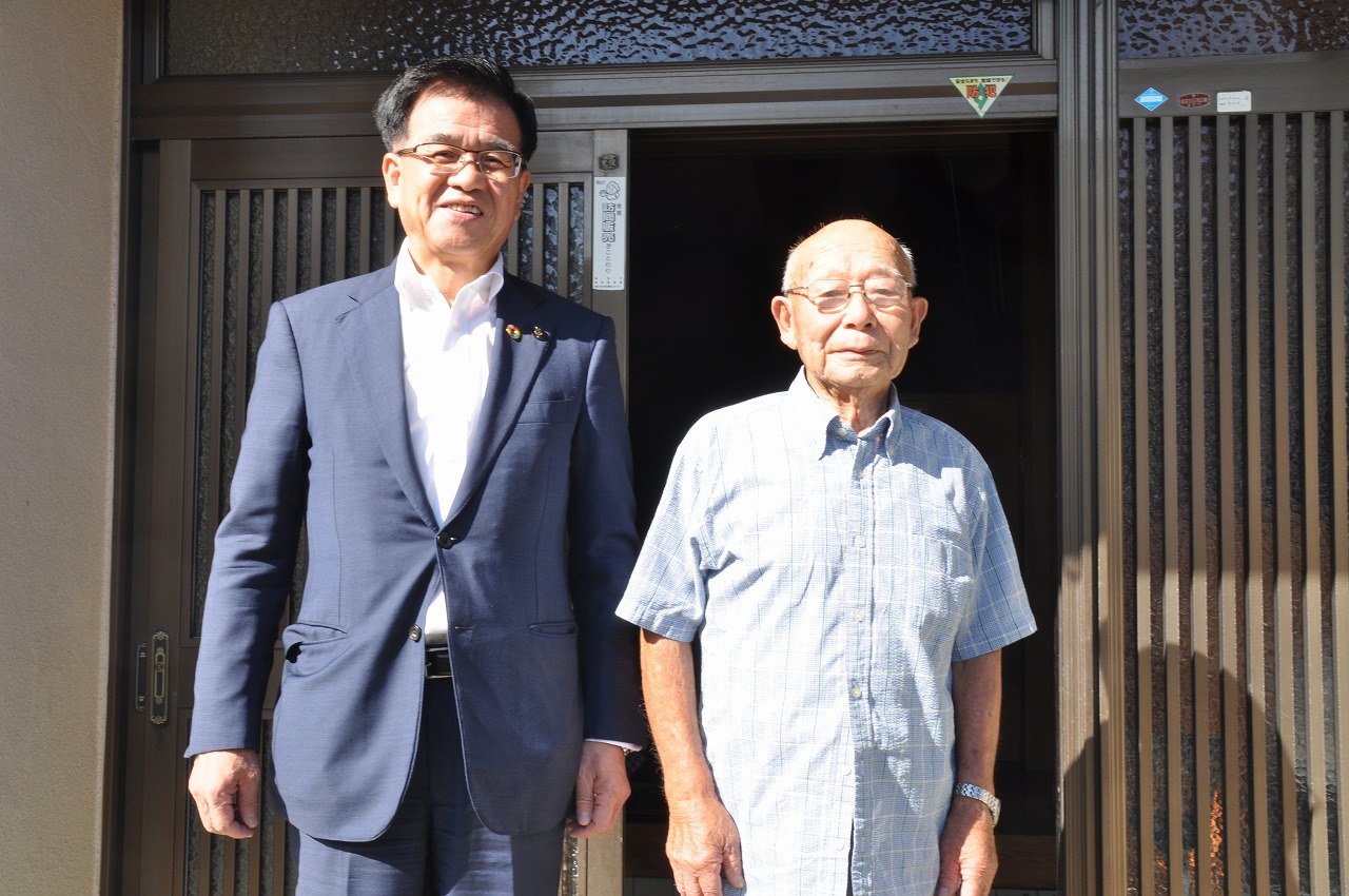 玄関前で、スーツを着た市長とチェック柄のシャツを着た高齢の男性が並び、カメラに向かって笑顔で記念撮影している写真