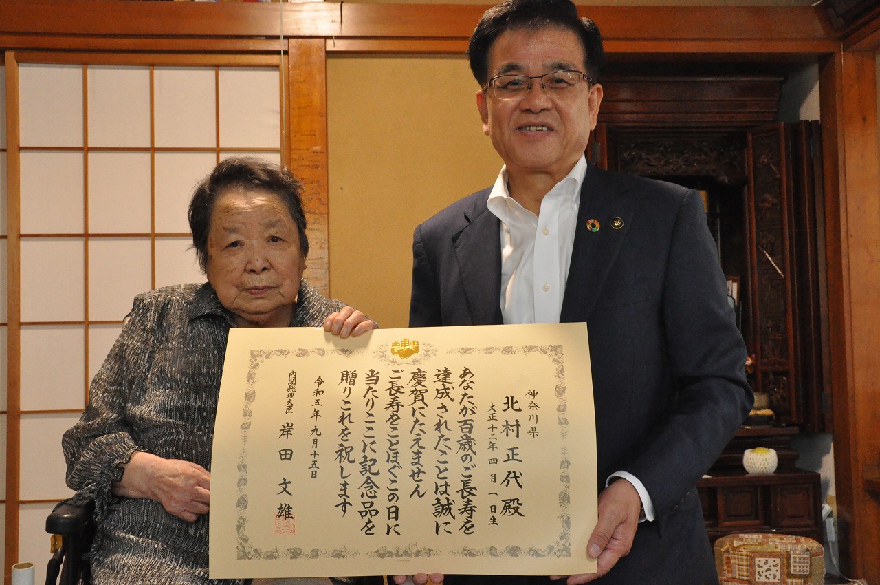 縁側のある和室で、スーツ姿の市長が賞状を高齢の女性と一緒に持ち笑顔で記念撮影している写真