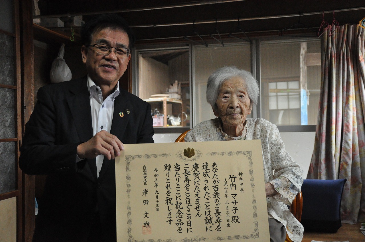 黒いスーツの市長と椅子に座った高齢女性が、部屋の中で賞状を広げて一緒に持ち記念撮影している写真