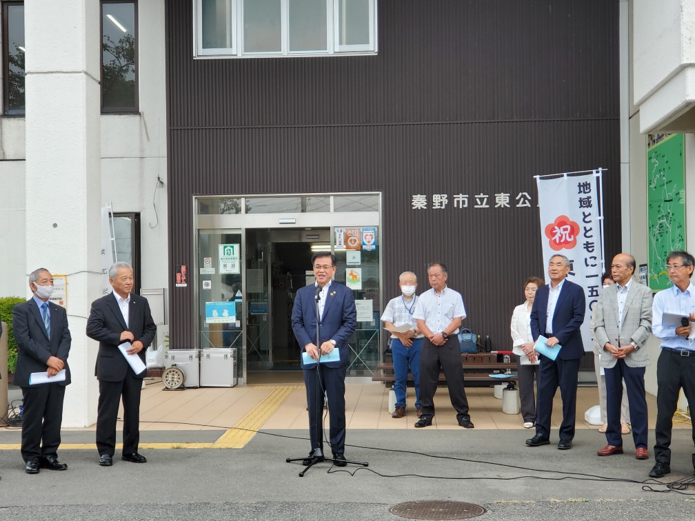 複数の男性が建物前に並び、中央の市長がマイクを持って式典の挨拶を行っている様子の写真