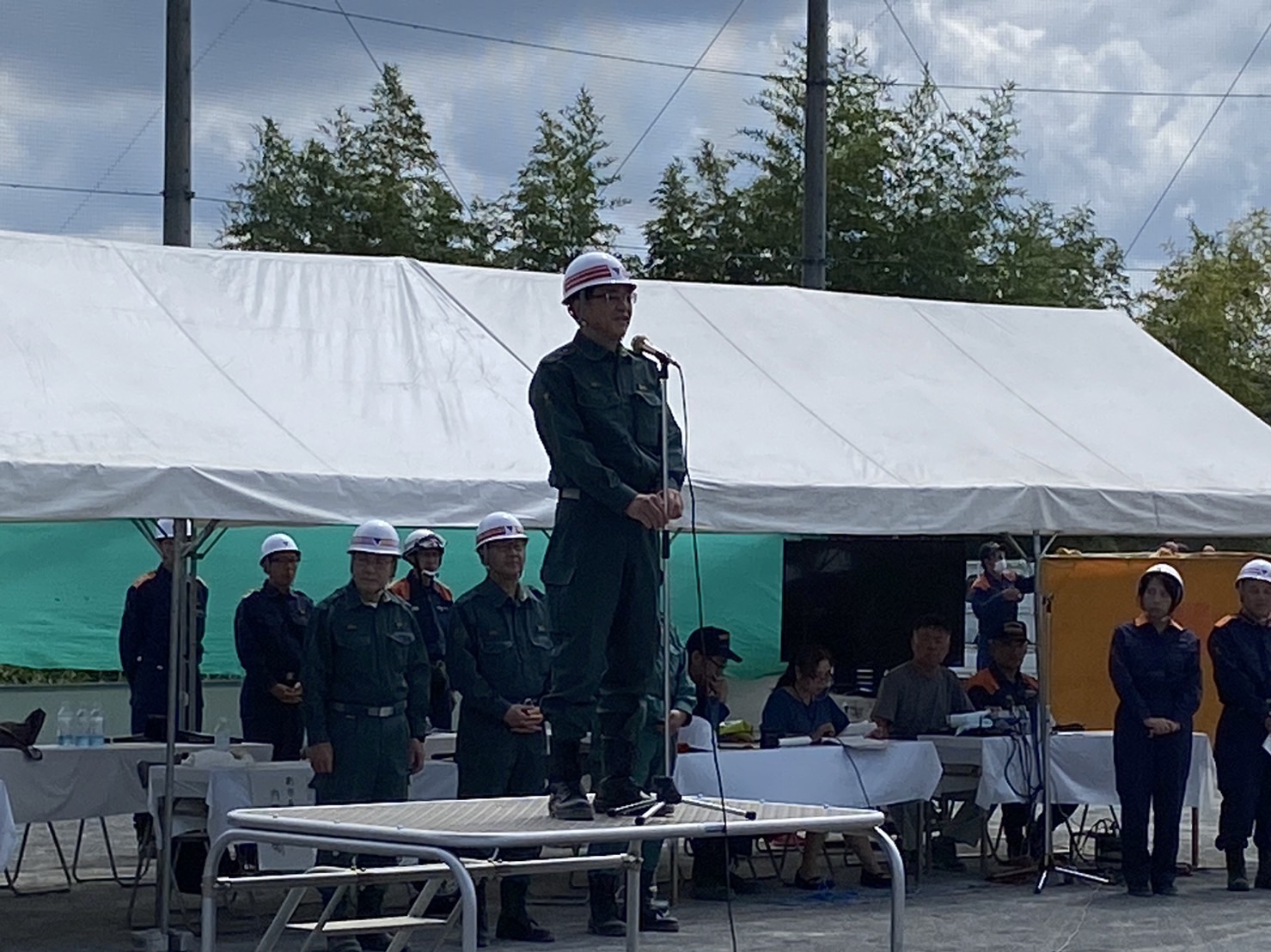 白いヘルメットと緑色の制服を着用した市長が、台の上からマイクでスピーチをしており、その後ろには同様の服装をした人々やテントが見える屋外イベントを写した写真