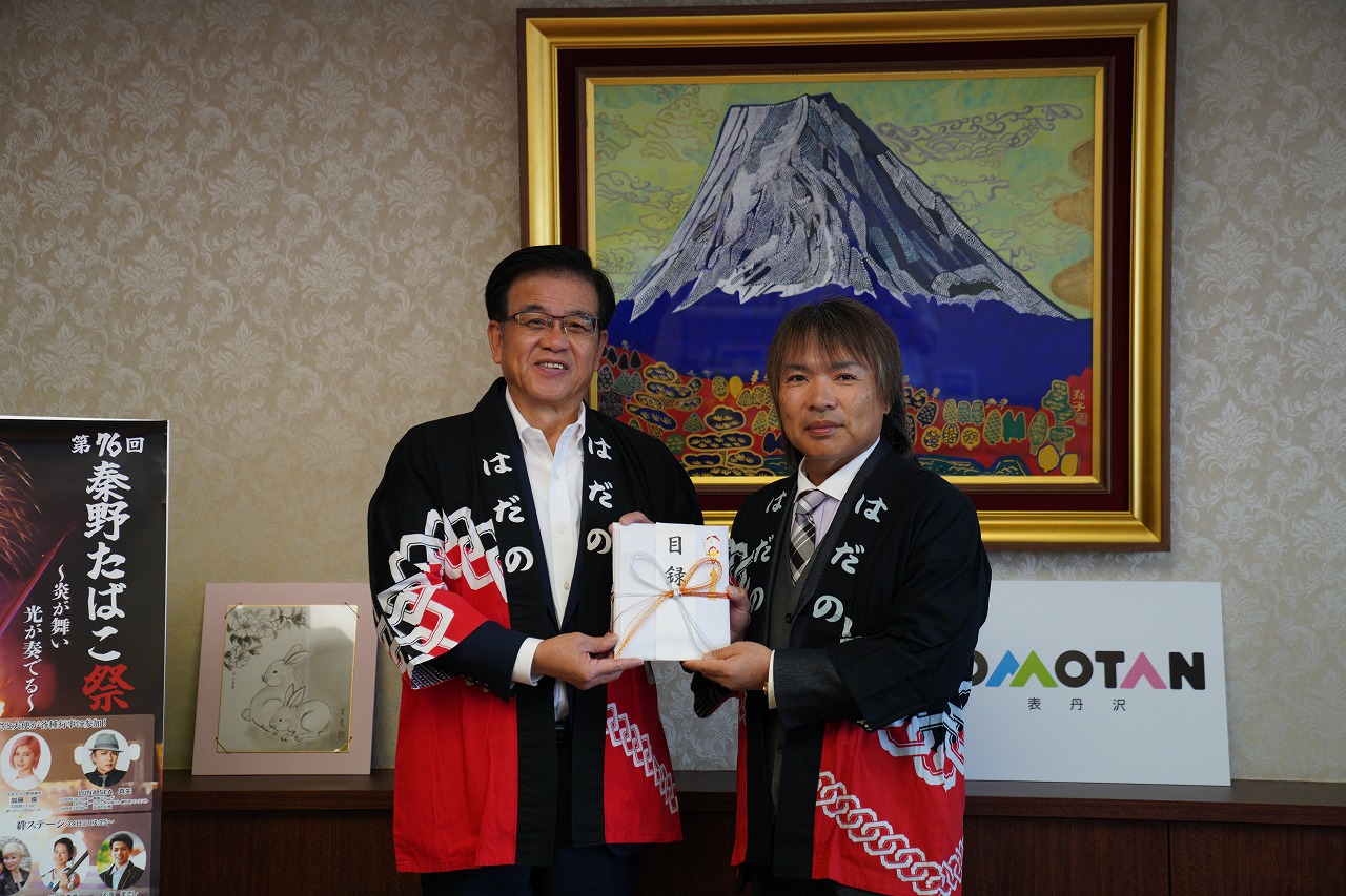 法被姿の市長とLUNA SEA真矢さん、目録と書かれた記念品を両手で一緒に持ちながら記念撮影している写真