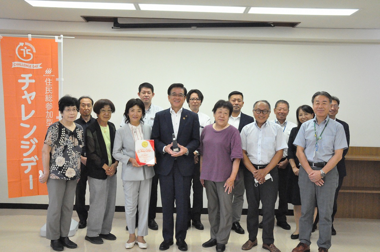 「チャレンジデー」と書かれたオレンジ色の旗の横で、スーツ姿の市長がトロフィーと賞状を持ち、様々な世代の男女十数名が並んでカメラに笑顔を向けている集合写真