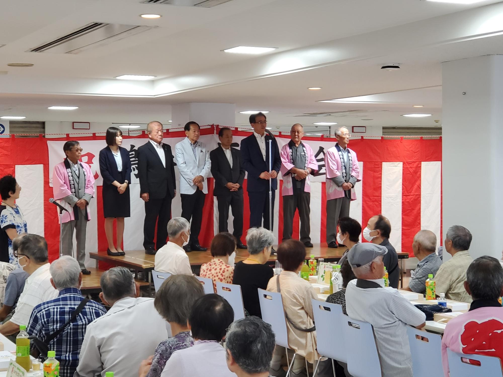 紅白の幕が張られたステージで、マイクを持つ市長を中心に法被を着た人やスーツ姿の人々が立ち、テーブル席に座る多くの聴衆がその様子を見ている様子の写真