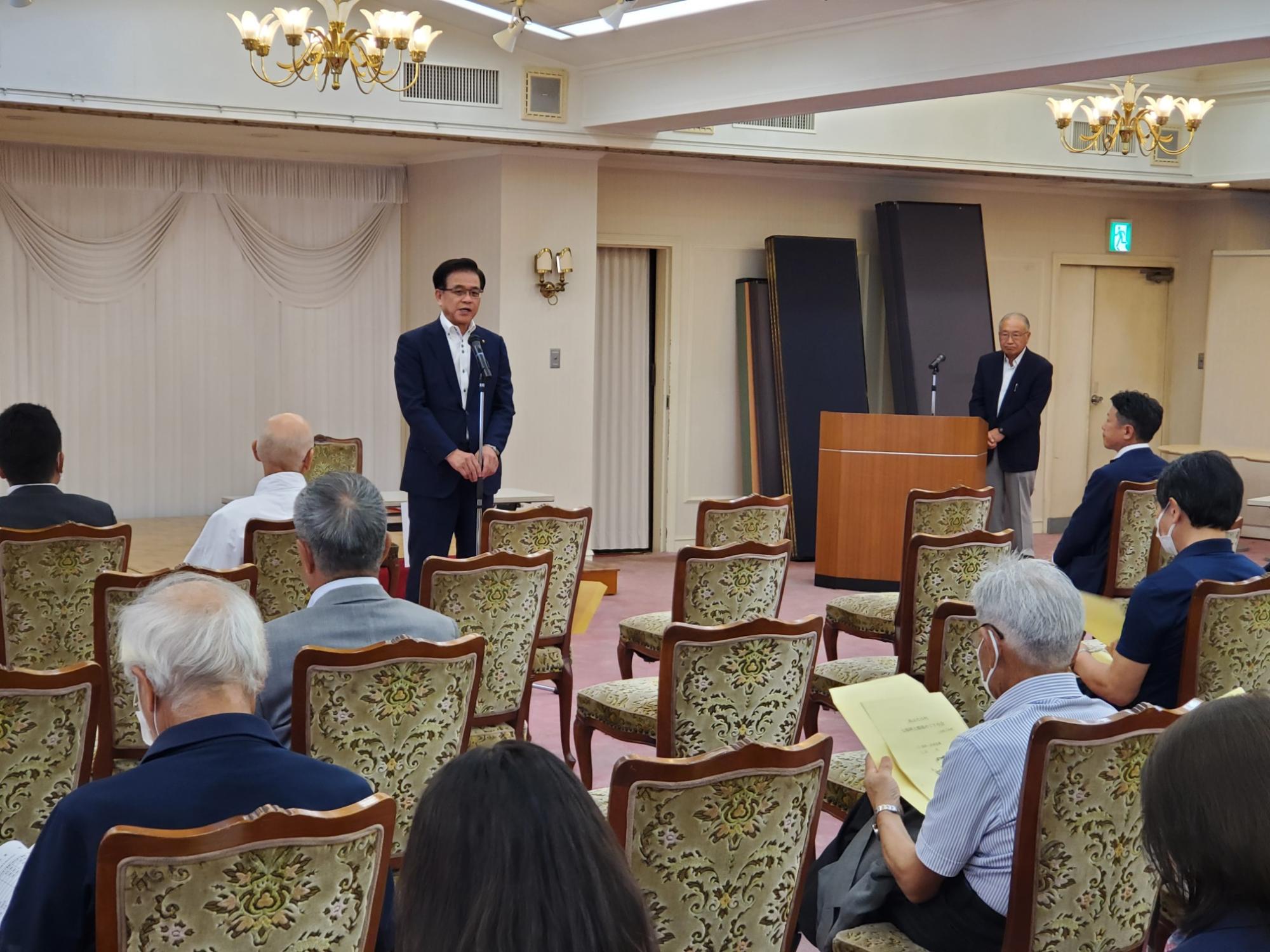 白い壁とシャンデリアのある室内で、スーツ姿の市長がマイクを使って聴衆に話しかけ、椅子に座った人々がその話を聞いている様子の写