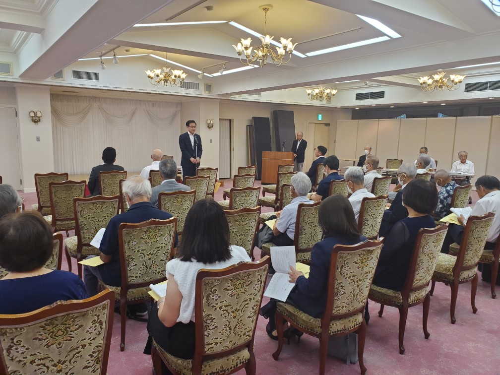 花柄の布張りの椅子が並んだ広い会場で、スーツ姿の市長が聴衆に向かって話す様子を後方から写した写真