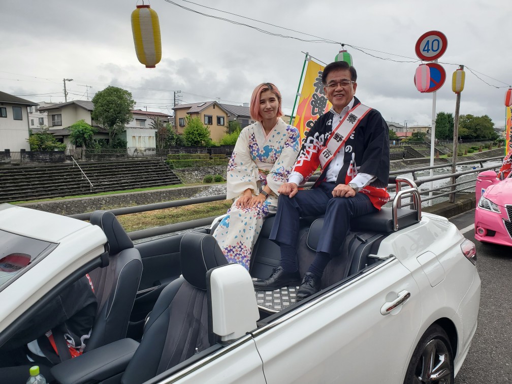 着物姿の若い女性と「野々市」と書かれたたすきをかけたスーツ姿の市長が、提灯やのぼりが飾られた川沿いの道路でオープンカーの後部座席に乗っている写真