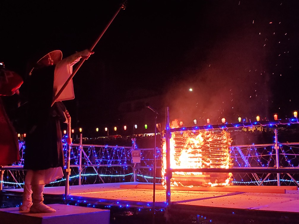 伝統的な衣装を着て編笠をかぶった人物が、火のついた櫓と青い電飾で飾られた祭りの船上で長い棒を掲げている写真