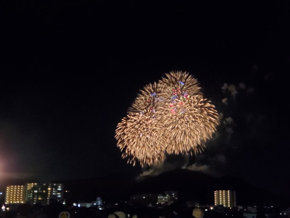 夏の夜空に金色と赤色の光が大きく花開いた花火が打ち上げられ、その下に街明かりと集まった人々が見える風景を撮影した写真