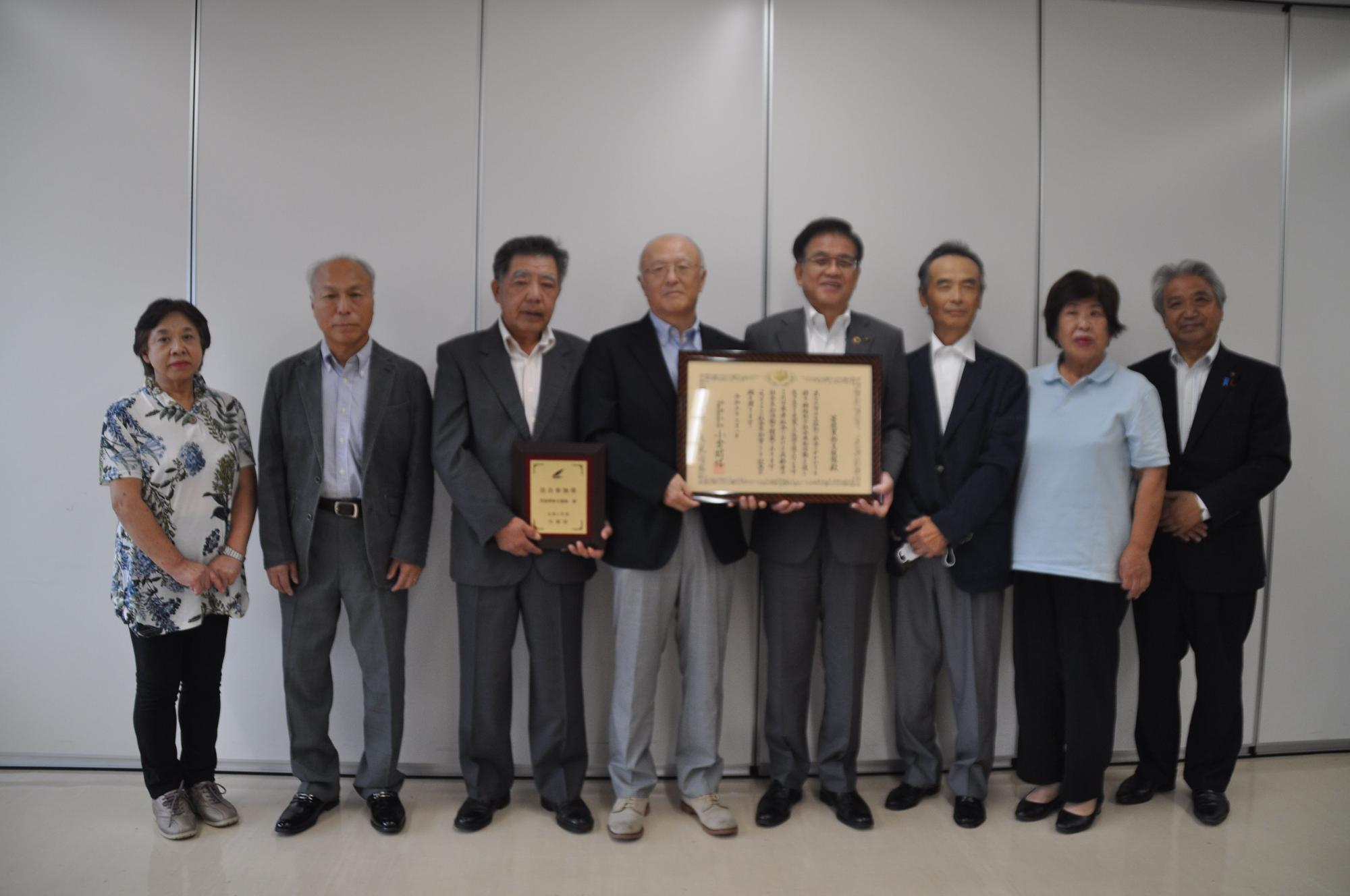 男性6人と女性2人が立ち並び、中央の男性と市長が額縁に入った大きな賞状や木製の盾を持っている集合写真