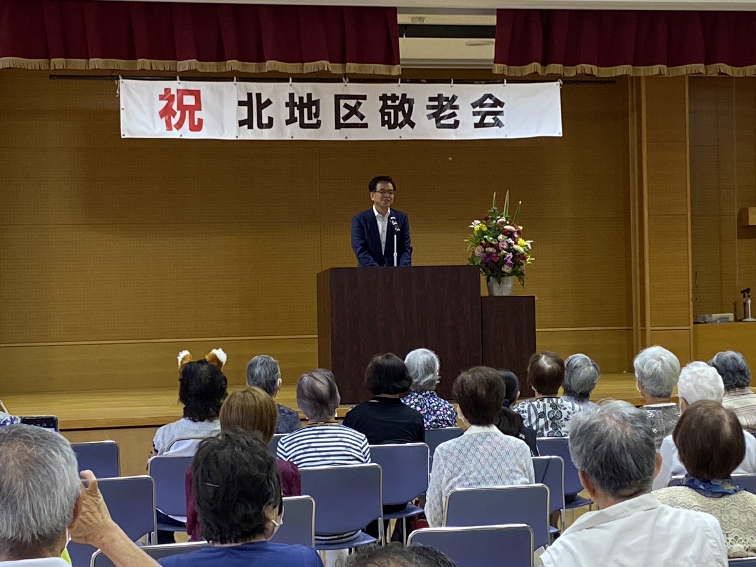 「祝 北地区敬老会」と書かれた看板が掲げられた舞台上で、スーツ姿の市長が演台から多くの参加者に向かって挨拶をしている様子の写真