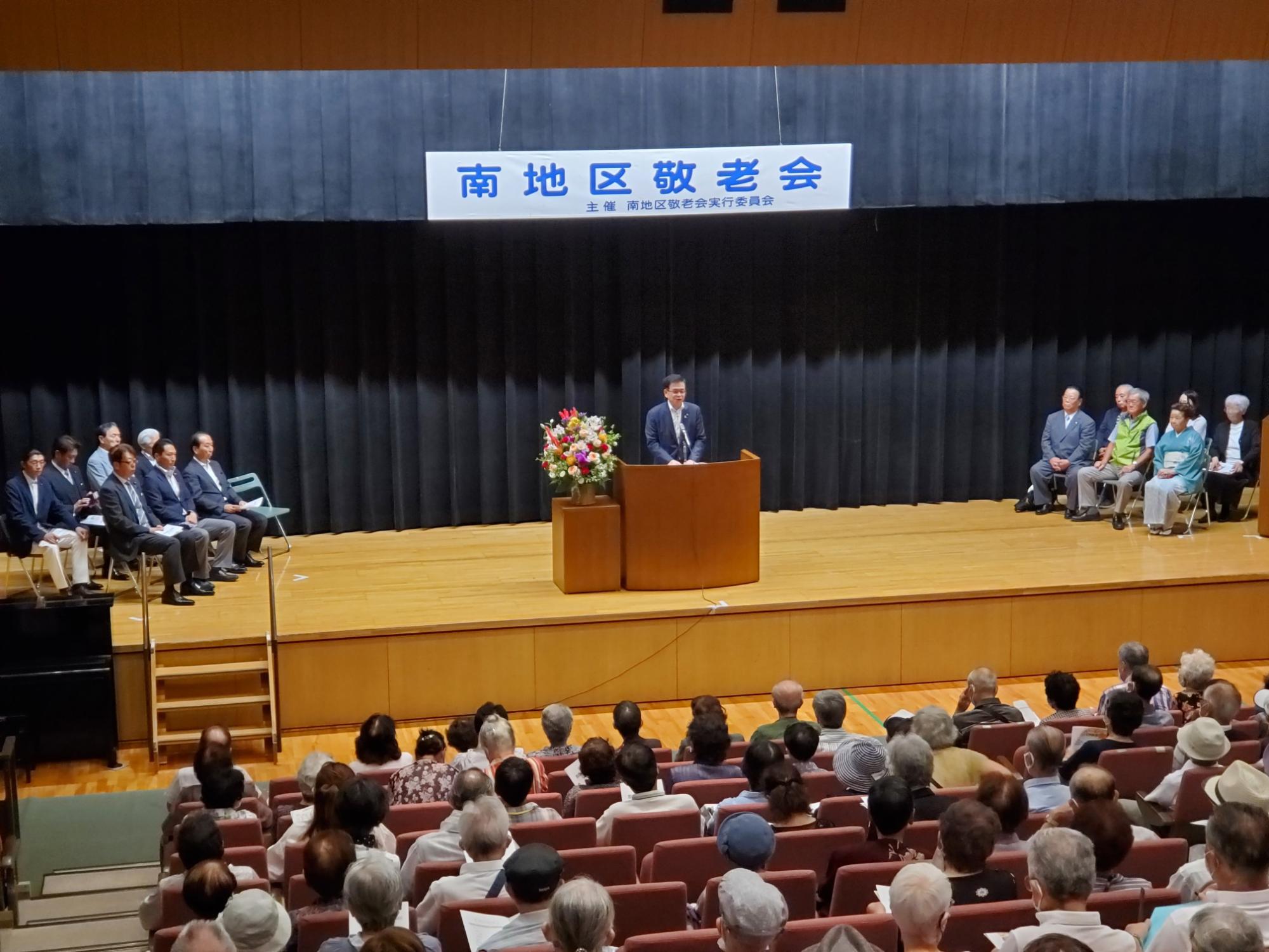 「南地区敬老会」と書かれた看板が掲げられた舞台で、マイクの前に立つ市長が聴衆に向けて話して様子を後方から写した写真