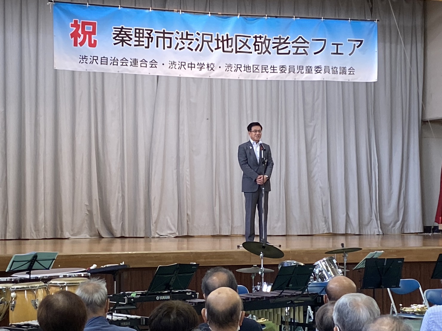 「秦野市渋沢地区敬老会フェア」と書かれた横断幕を背景に、ステージ上でマイクスタンドを前に話をする高橋市長を客席側から見た写真