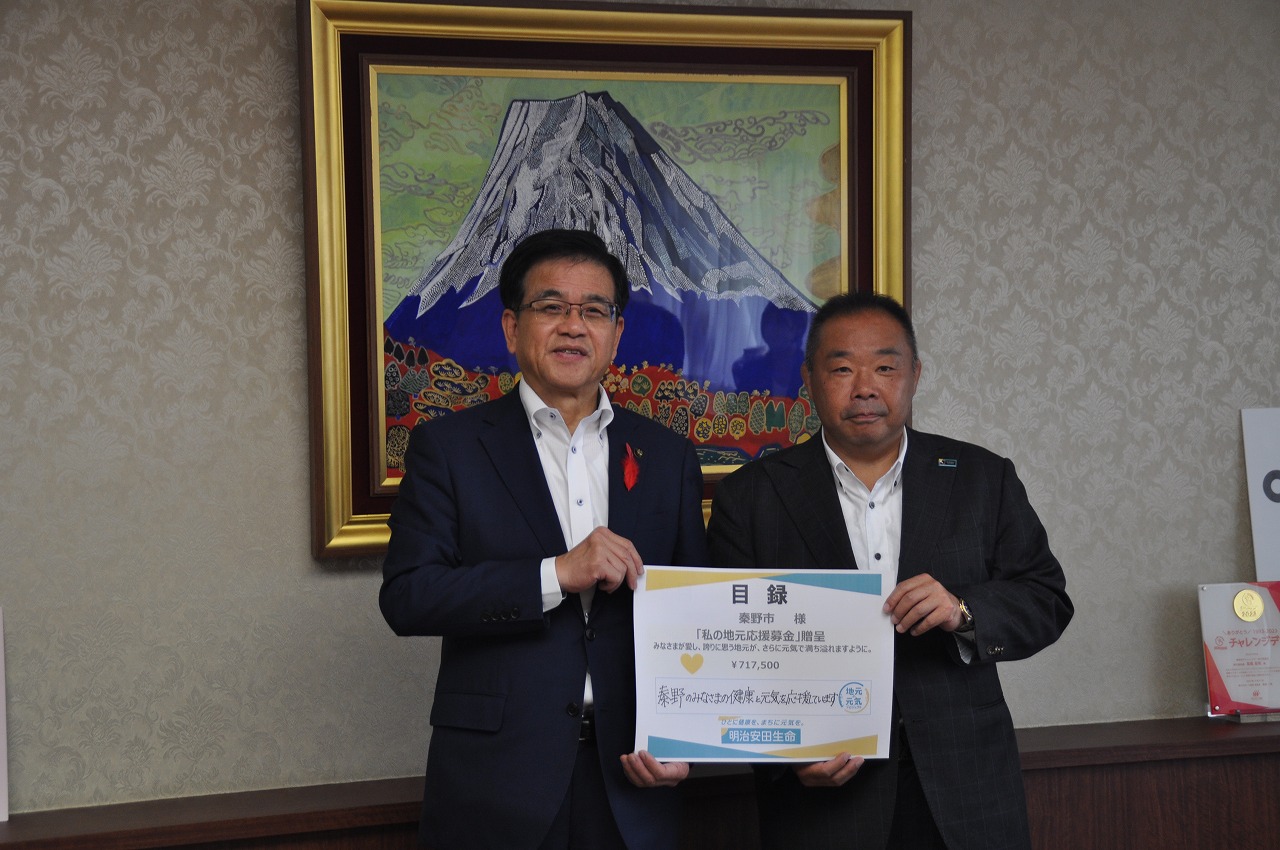 応接室のような部屋で、寄付の目録を一緒に持っている高橋市長と男性を正面から撮影した写真