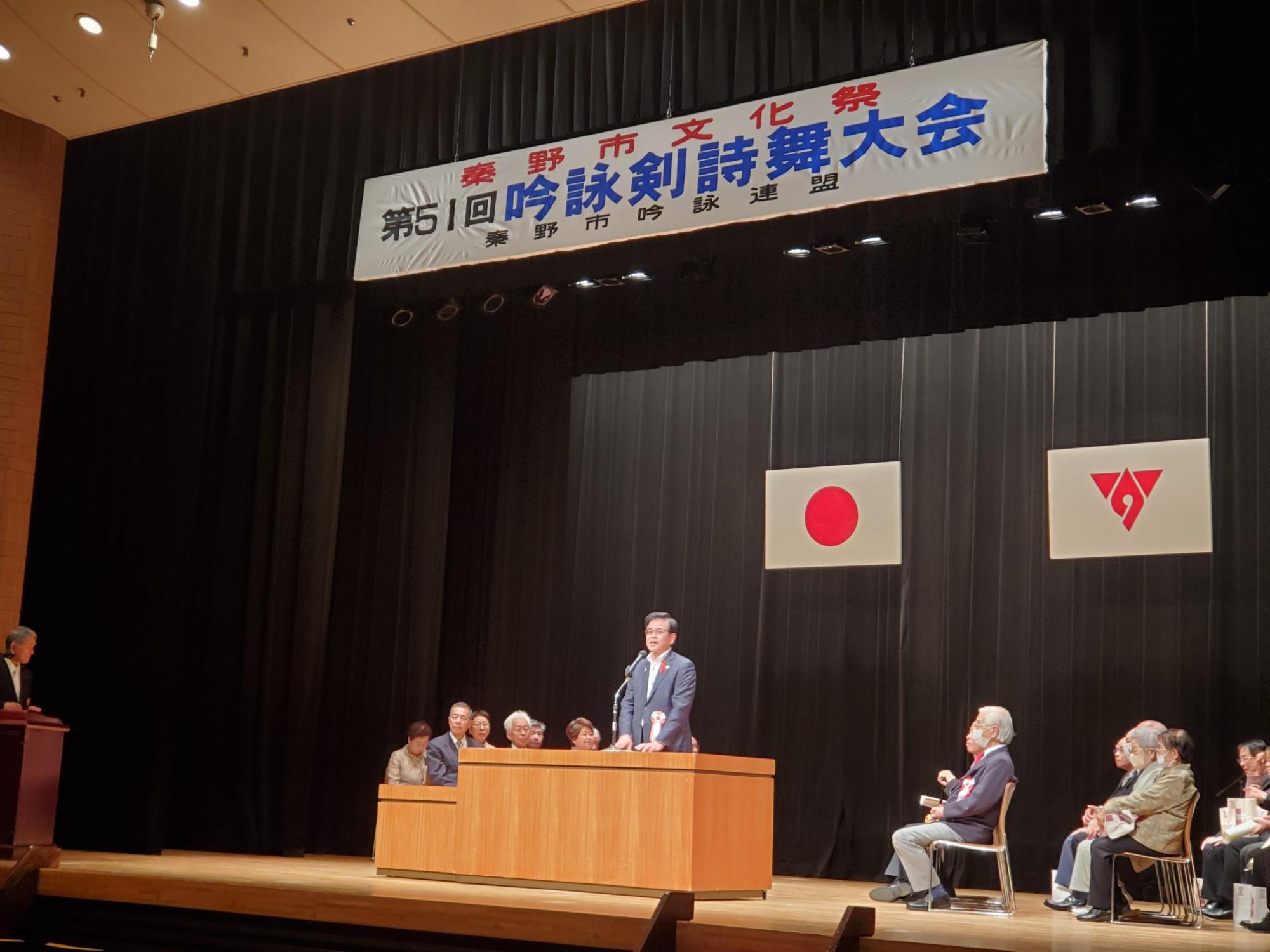 「秦野市文化祭吟詠剣詩舞大会」の横断幕の下、ステージの壇上でマイクスタンドに向かってスピーチをする高橋市長を捉えた写真