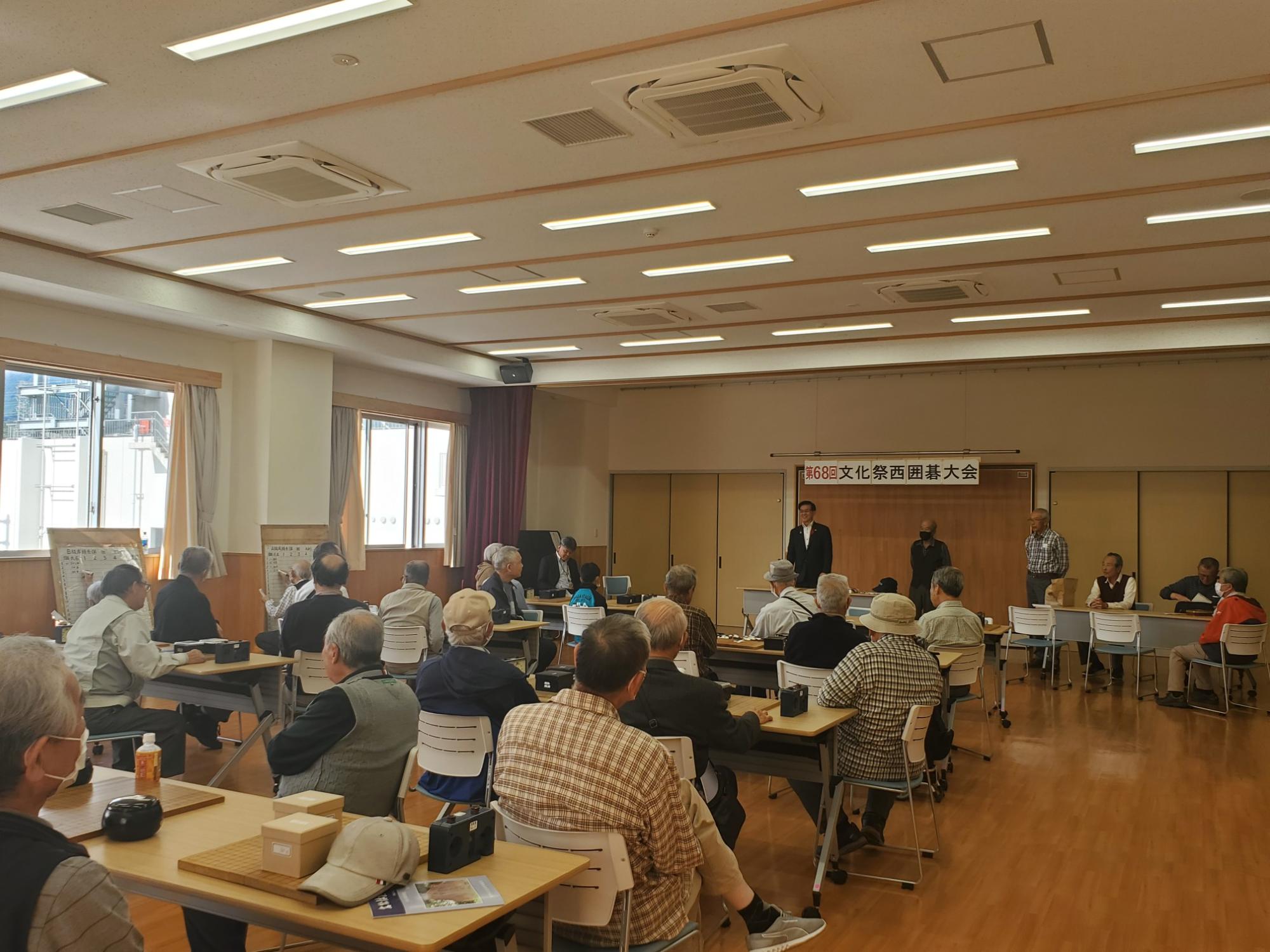 広い部屋の一室で、高橋市長と男性が立っており、囲碁が机の上に置かれて座る複数の参加者の様子を後ろから捉えた写真