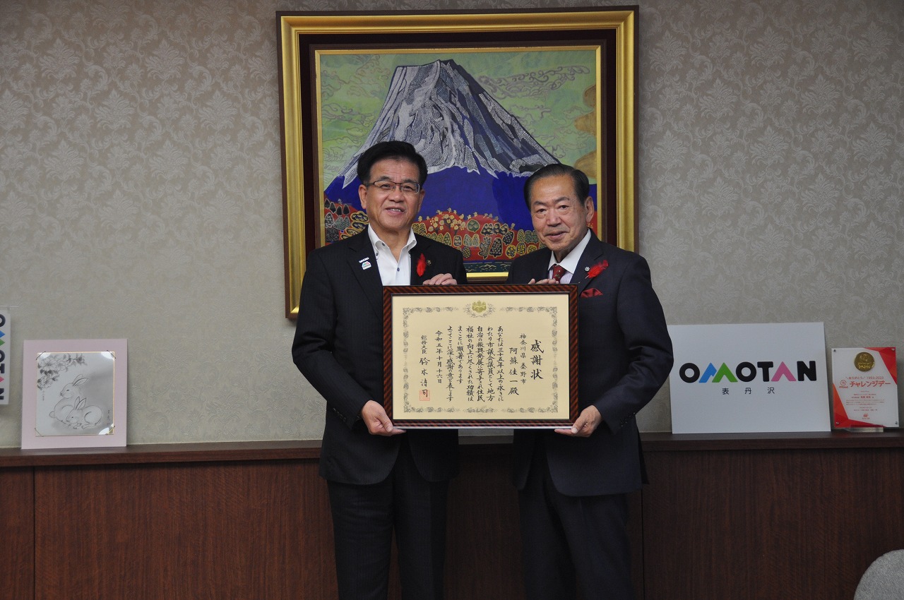 応接室のような室内で、高橋市長と男性が賞状の額縁を両手で持ち、並んで笑顔で立っている写真