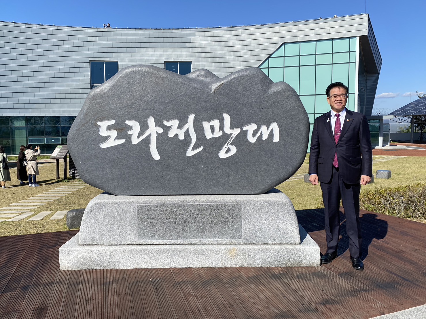 青空が広がる空の下、建物を背景に、ハングル文字で刻まれた大きな石碑の横で、高橋市長が笑顔で立っている写真