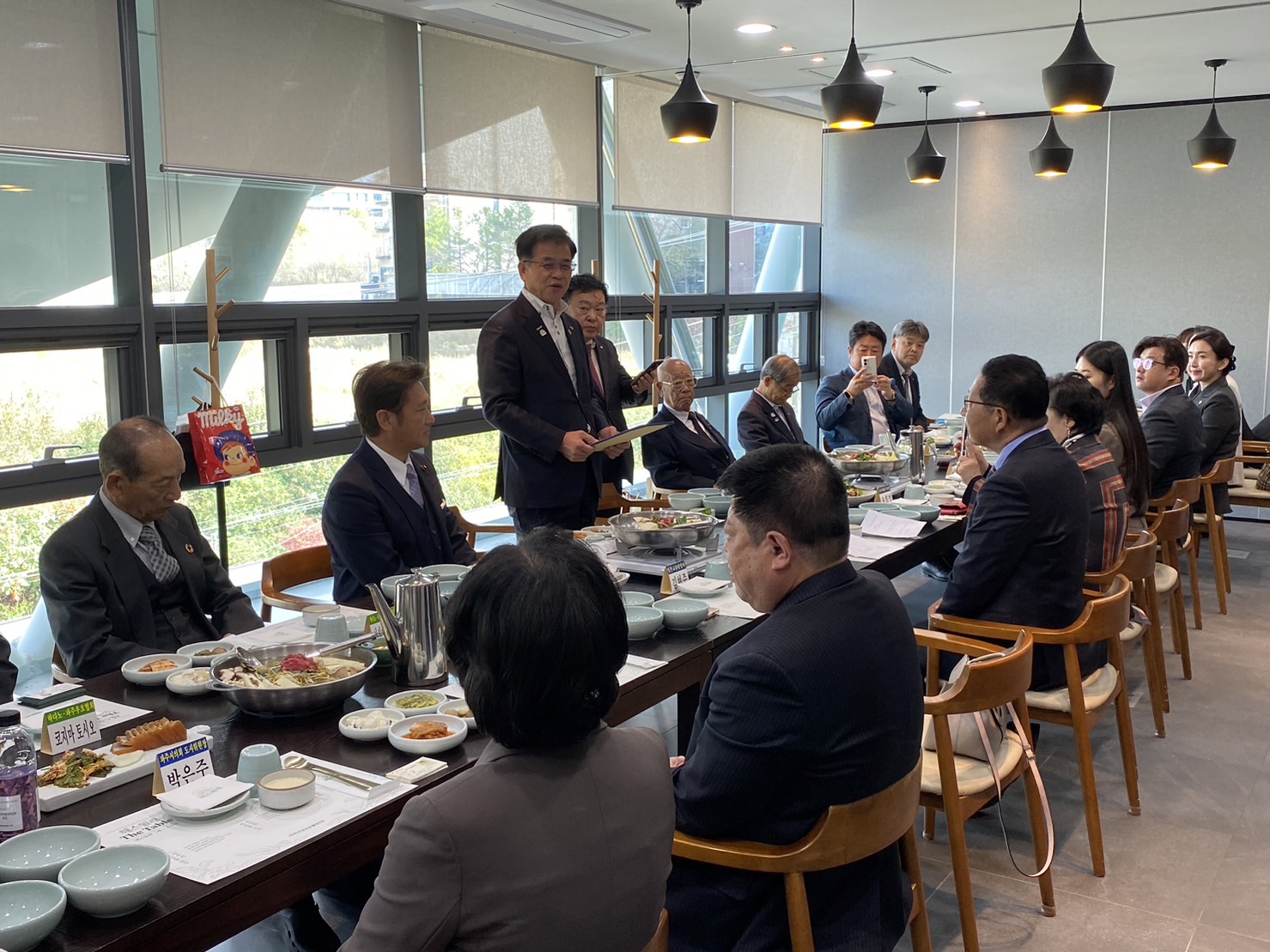 長机を囲んで座る多くの人々が食事をしているレストランのような部屋で、高橋市長が立って話している様子を捉えた写真