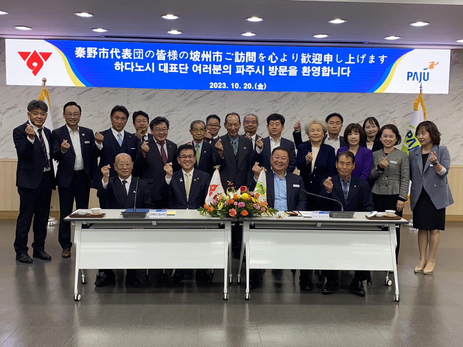 「秦野市代表団の皆様の坡州市ご訪問を心より歓迎申し上げます」と書かれた横断幕を背景に、老若男女の関係者と高橋市長が親指と人差し指をクロスしたポーズで記念撮影している写真