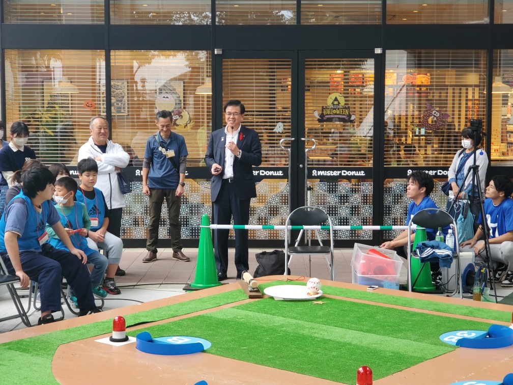 三角コーンや人工芝が置かれた屋外で、椅子に座っている複数の参加者の前で、話をする高橋市長の写真