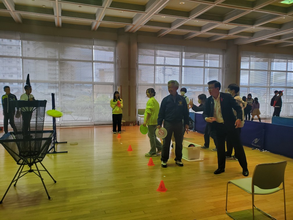 室内の広いフロアで、ディスクゴルフ用のゴールとコーンが設置されており、高橋市長がフリスビーを持ち、複数の人々がそれを見ている写真