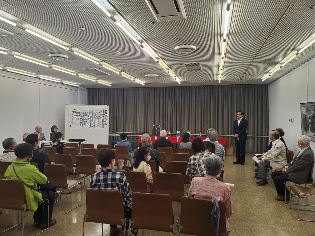 講堂のような部屋で、様々な人々が席に座っており、高橋市長が立って話をしている様子の写真