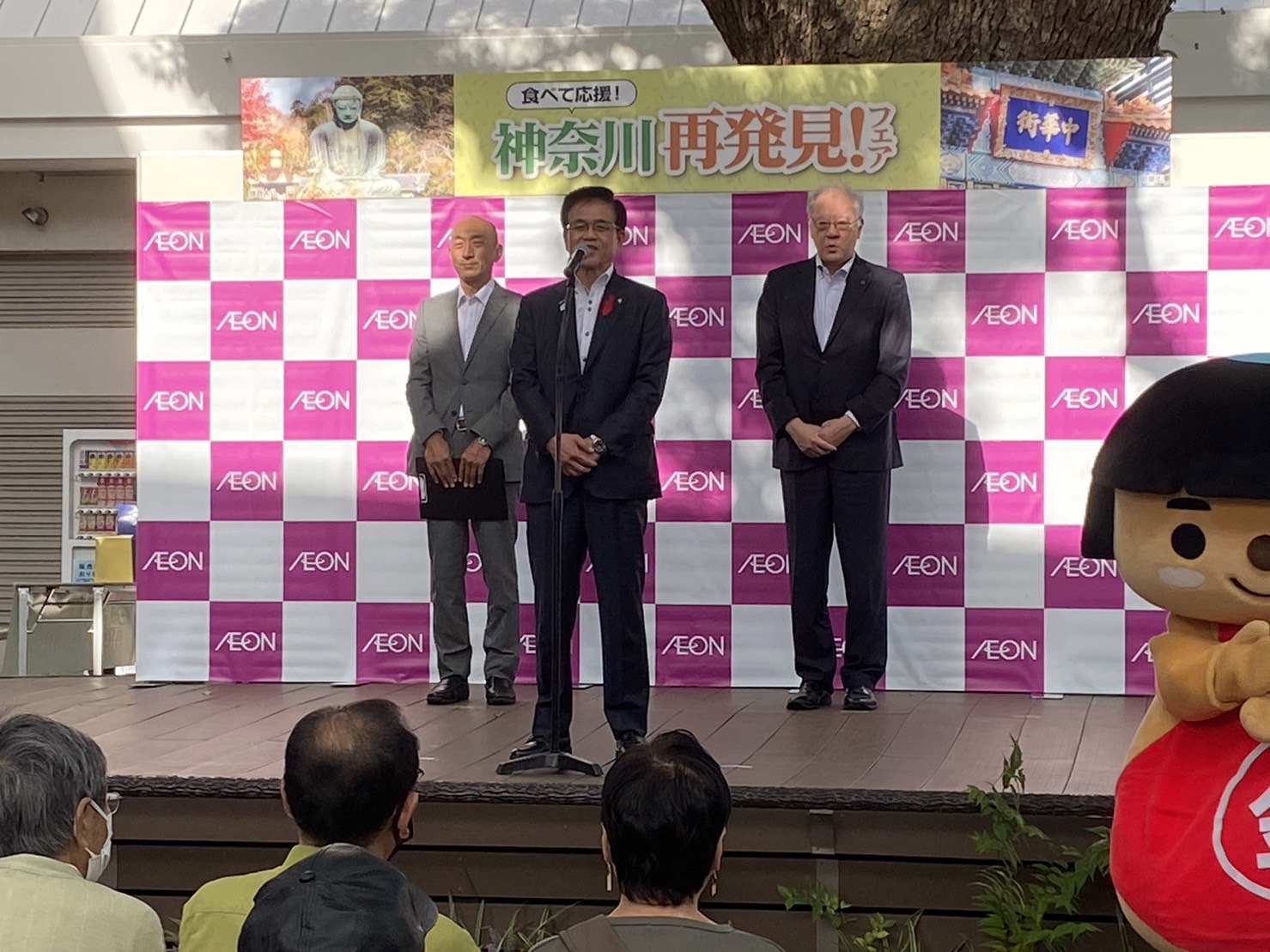 「AEON」のロゴが入ったピンクと白のチェック柄のバックパネル前で、男性2人とマイクスタンドの前で話をする高橋市長の写真