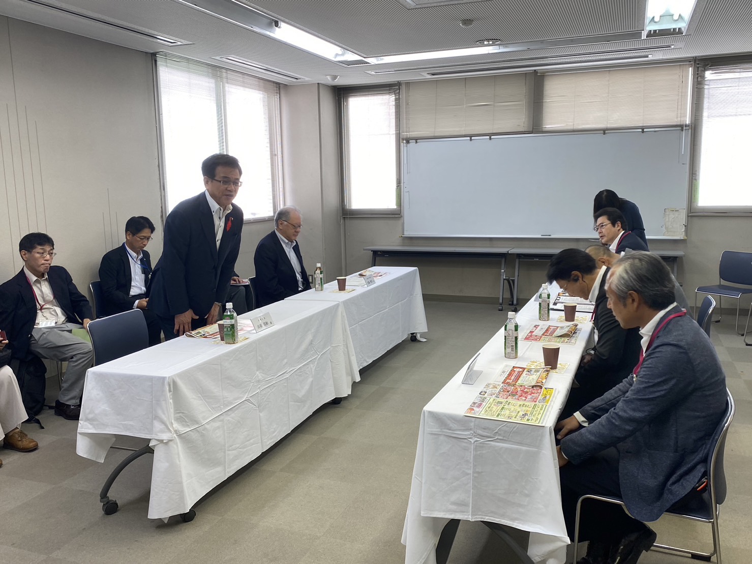 向かい合わせに配置された会議机で、高橋市長が立って話をしている様子を捉えた写真