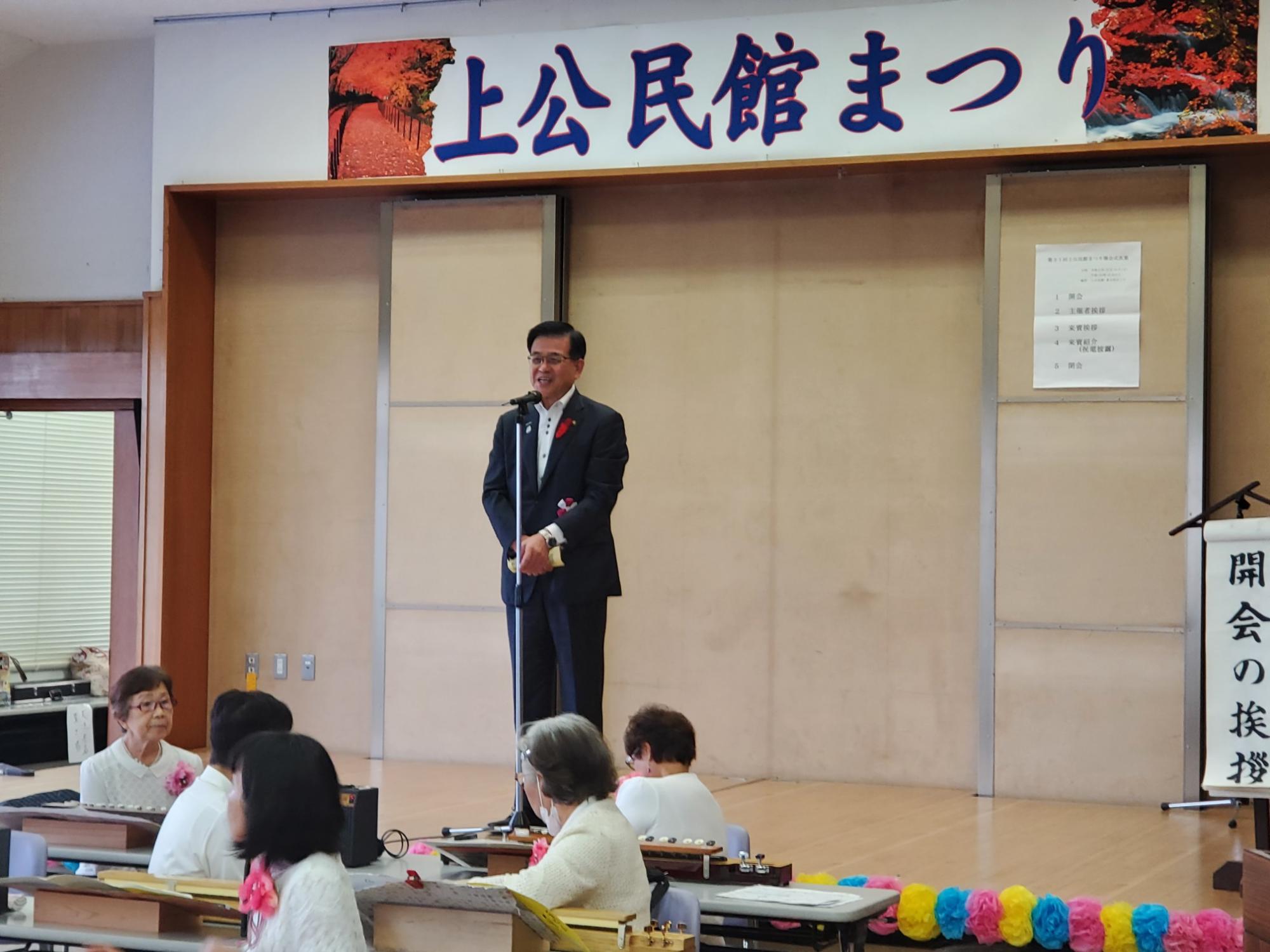 「上公民館まつり」と書かれた横断幕が掲げられたステージ上で、マイクを持って話す高橋市長を、着席した複数の観客が聞いている屋内の写真