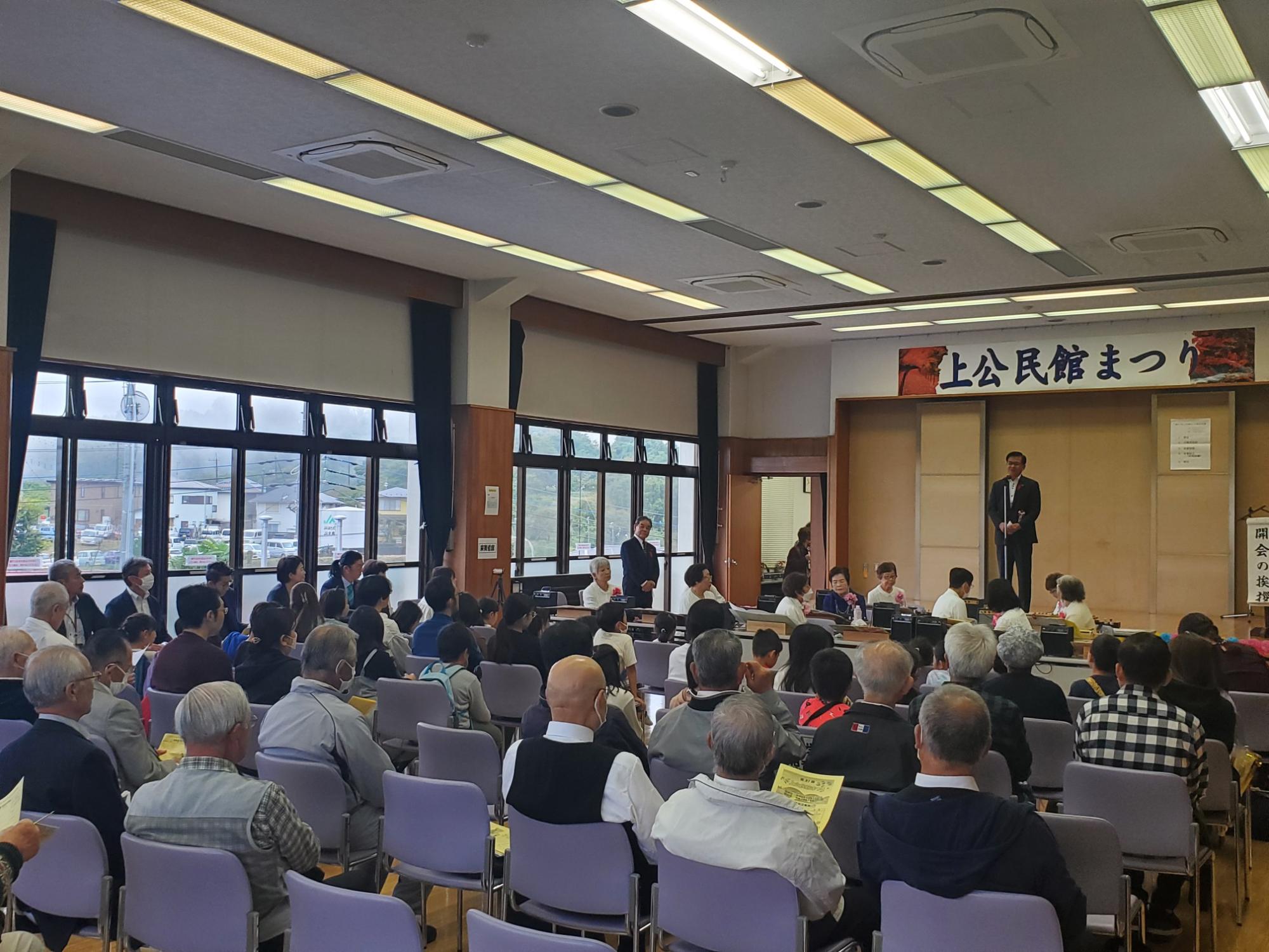 「上公民館まつり」と書かれた横断幕の下、大勢の参加者が椅子に座っている会場で、マイクを持って話す高橋市長を後方から捉えた写真