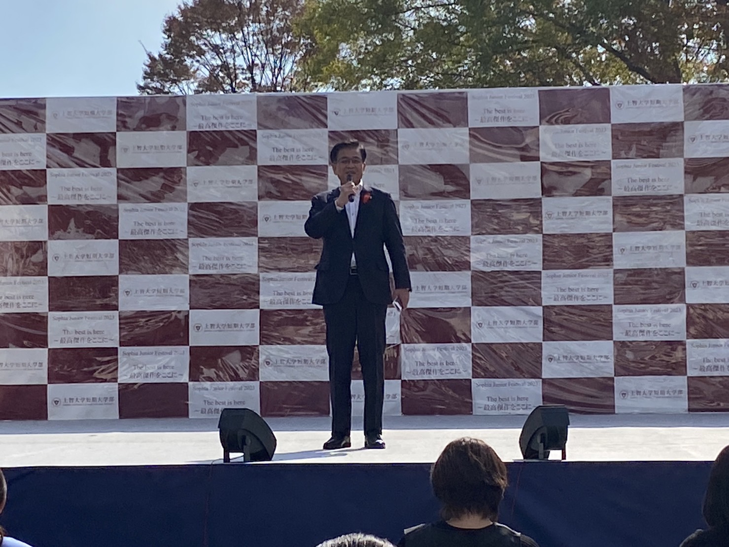 あずき色のバックパネルを背景に、野外ステージでマイクを持って話をする高橋市長の写真