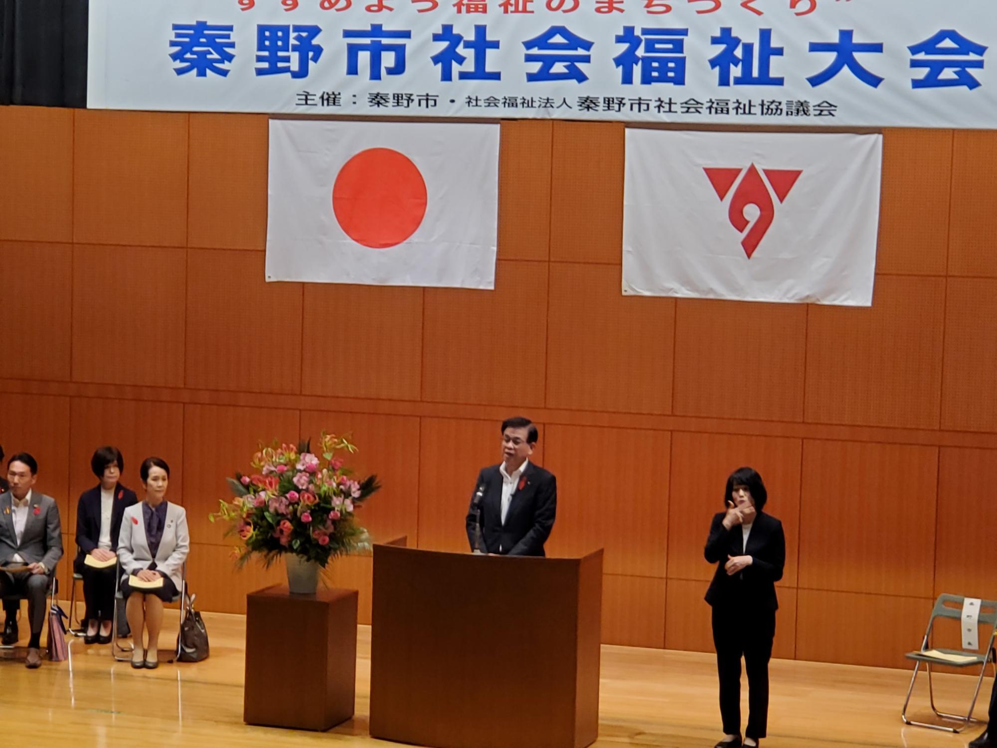 「秦野市社会福祉大会」と書かれた横断幕と、日本の国旗、秦野市の国旗が掲げられ、壇上でスピーチをする高橋市長の隣で女性が手話通訳をしている様子の写真