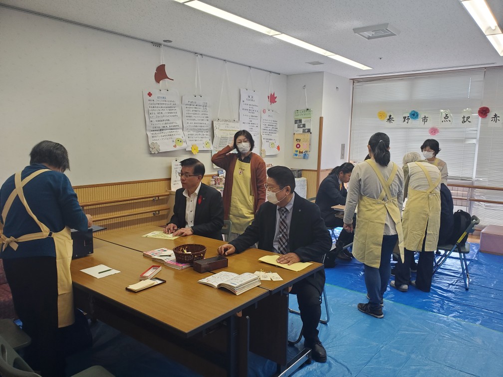 青いビニールシートが床に敷かれた室内で、高橋市長と関係者がの男性が机に向かって座っており、周りには黄色のエプロン姿の女性たちが複数立っている写真