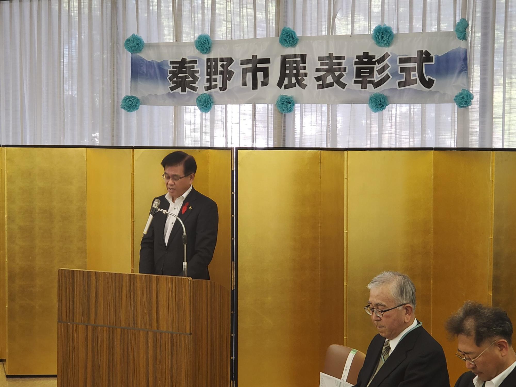 「秦野市展表彰式」の横断幕があり、金屏風が設置されたステージの壇上で、マイクに向かってスピーチをする高橋市長の写真