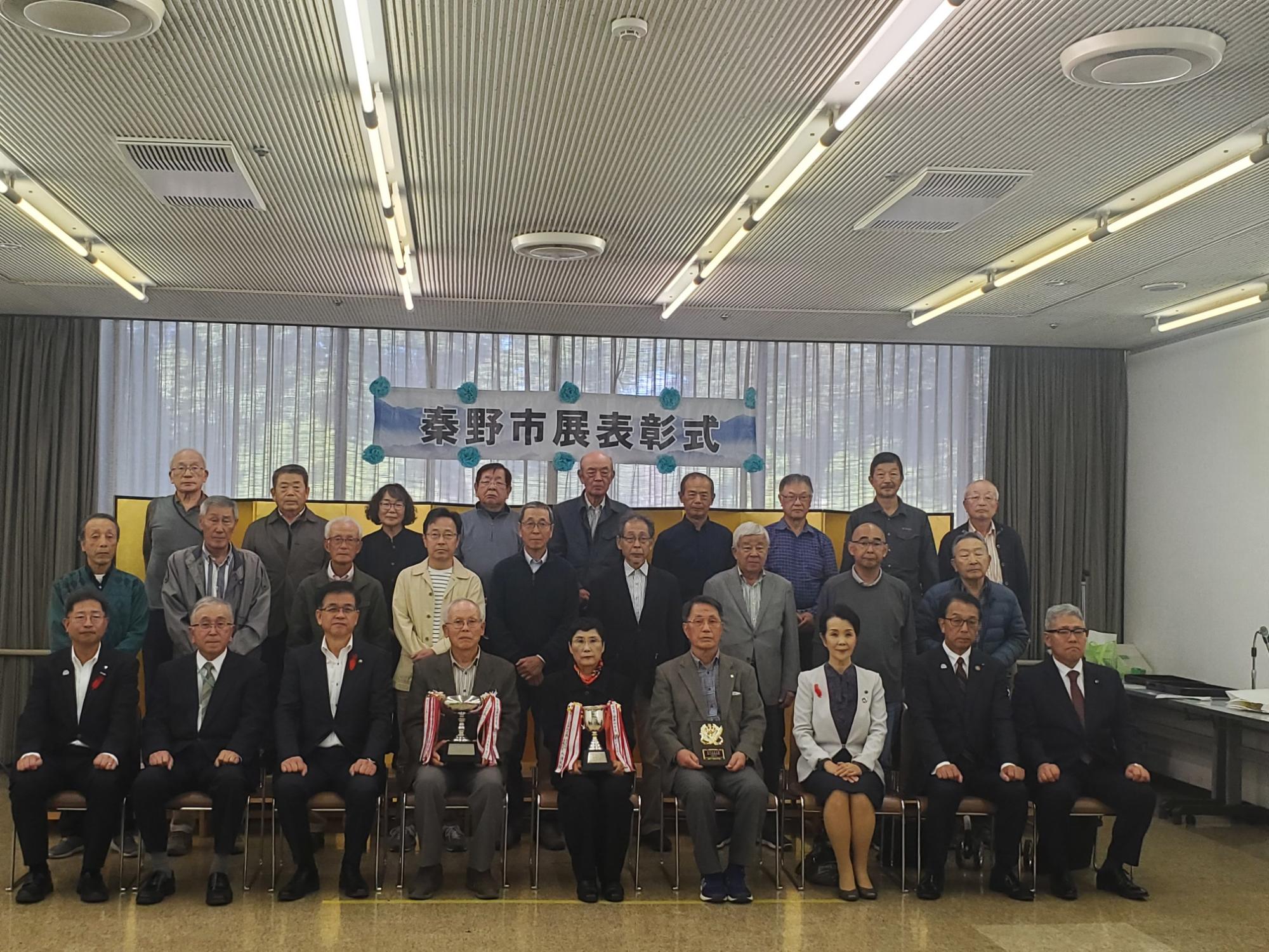 「秦野市展表彰式」の横断幕があり、トロフィーや盾を手にした30名ほどの男女と高橋市長が記念撮影している写真