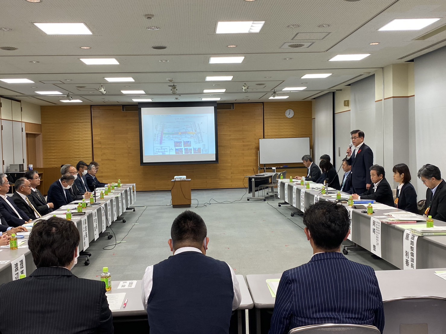 会議室の中央に置かれたスクリーンに資料が投影され、複数の参加者がU字型のテーブルに座り、壇上に立つ高橋市長の話を聞いている様子の写真