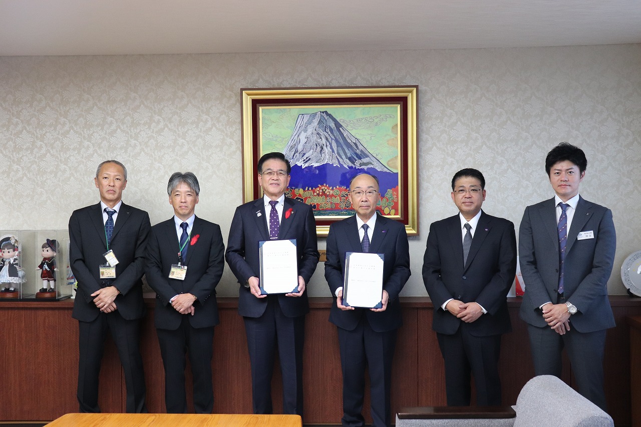 応接室のような部屋で、協定書のようなものを高橋市長と関係者が手に持ち、関係者4名の男性が並んでいる写真