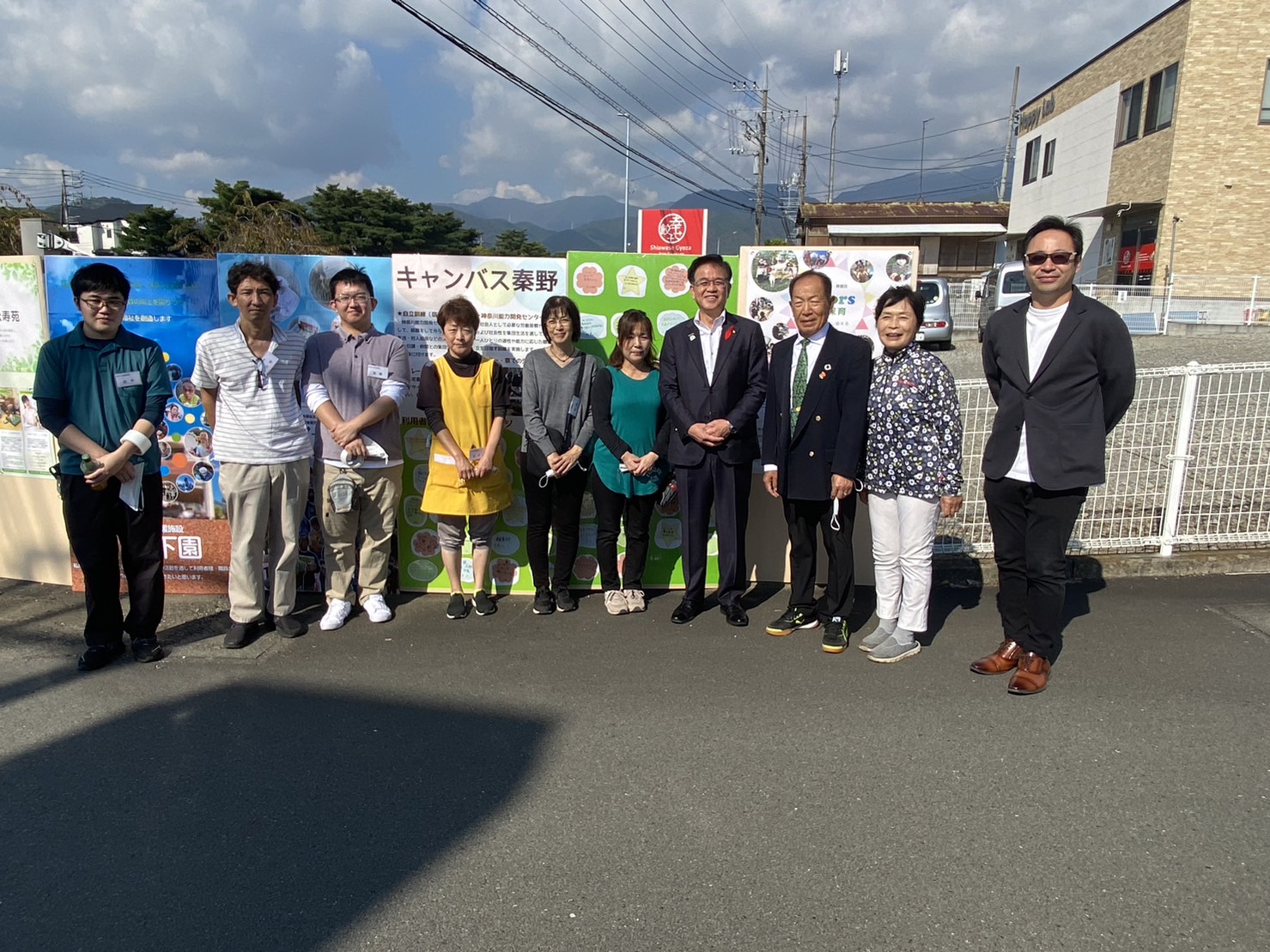 広々とした駐車場のような屋外で、高橋市長と複数の男女が横一列に並び記念撮影している写真