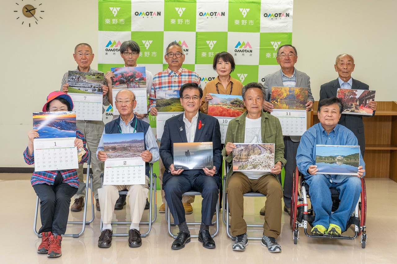 「秦野市」と「OMOTAN」のロゴが入ったバックパネル前で、高橋市長と複数の人々が写真付きカレンダーを持っている合写真