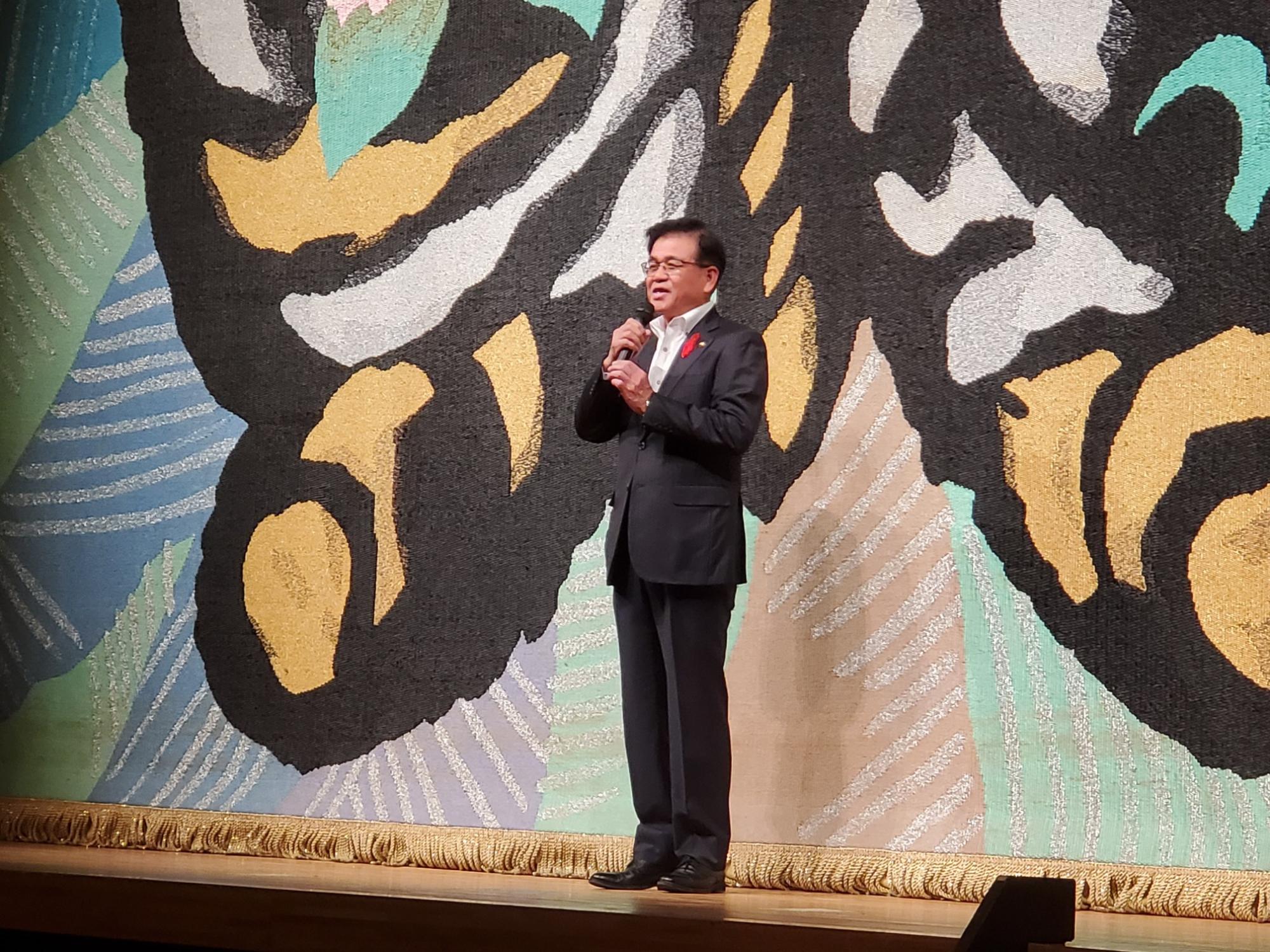 オレンジ薄緑色を基調としたで抽象的な模様の緞帳の前で、マイクを持ちスピーチをしている高橋市長の写真