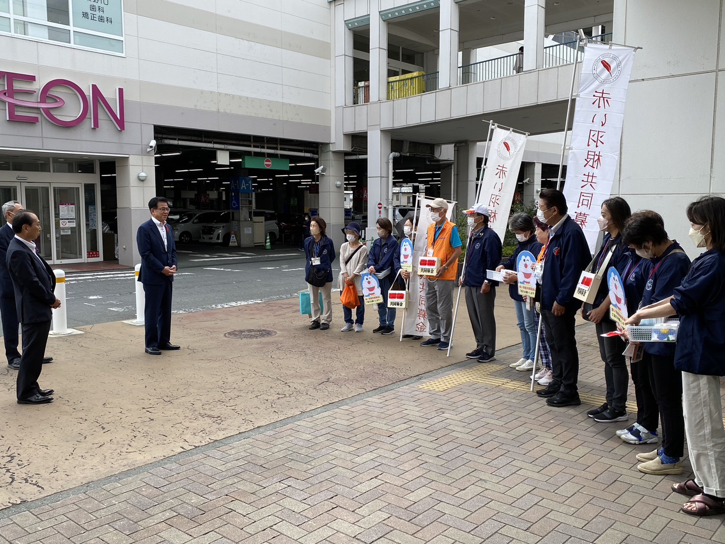 イオンの建物前で、高橋市長がが横一列に並んで募金箱や旗を持っている赤い羽根共同募金活動をしているグループに向けて話をしている写真