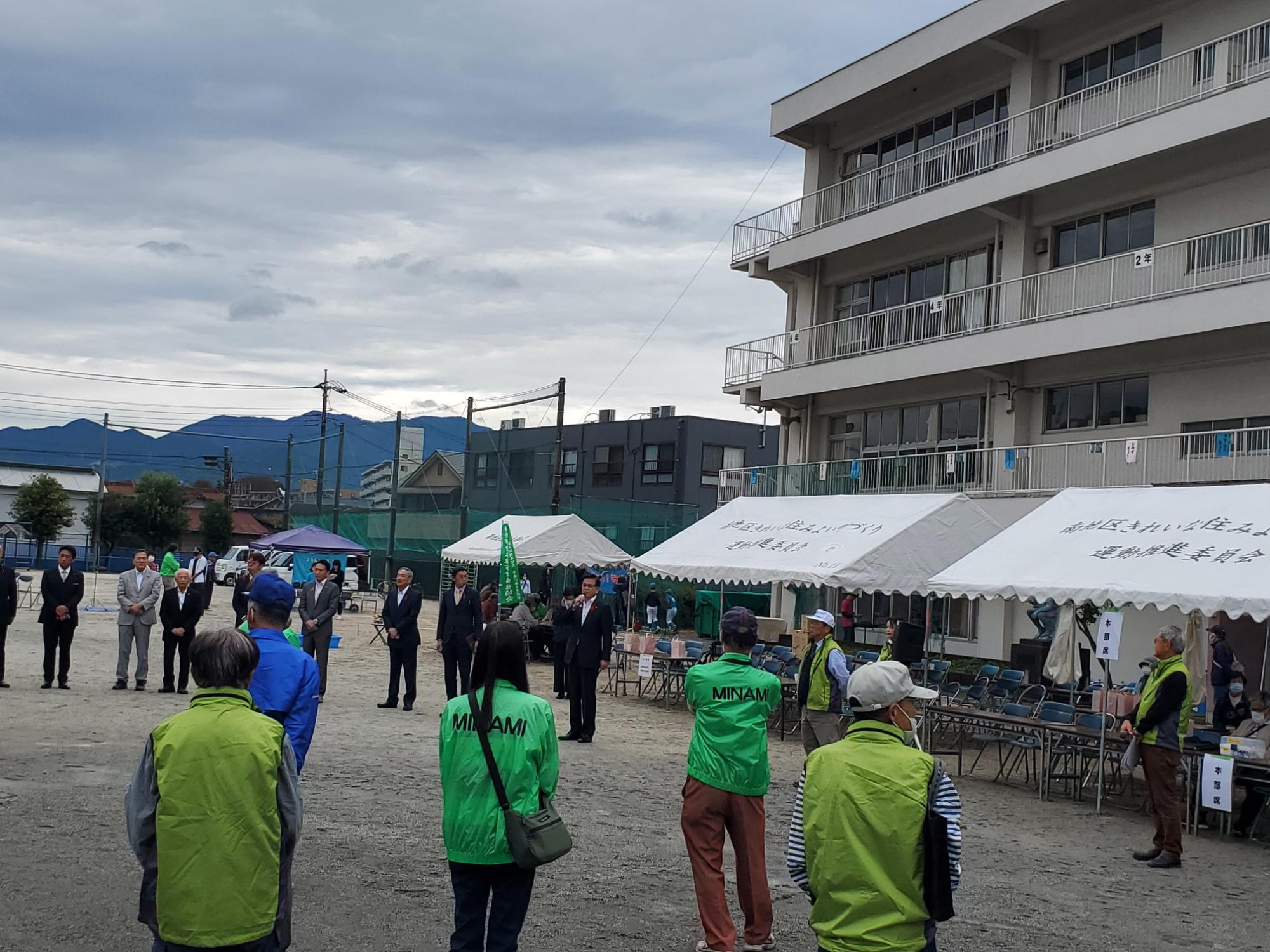 テントが複数設置されている屋外の広場で、高橋市長が話をしており、関係者がその話を聞いている様子の写真