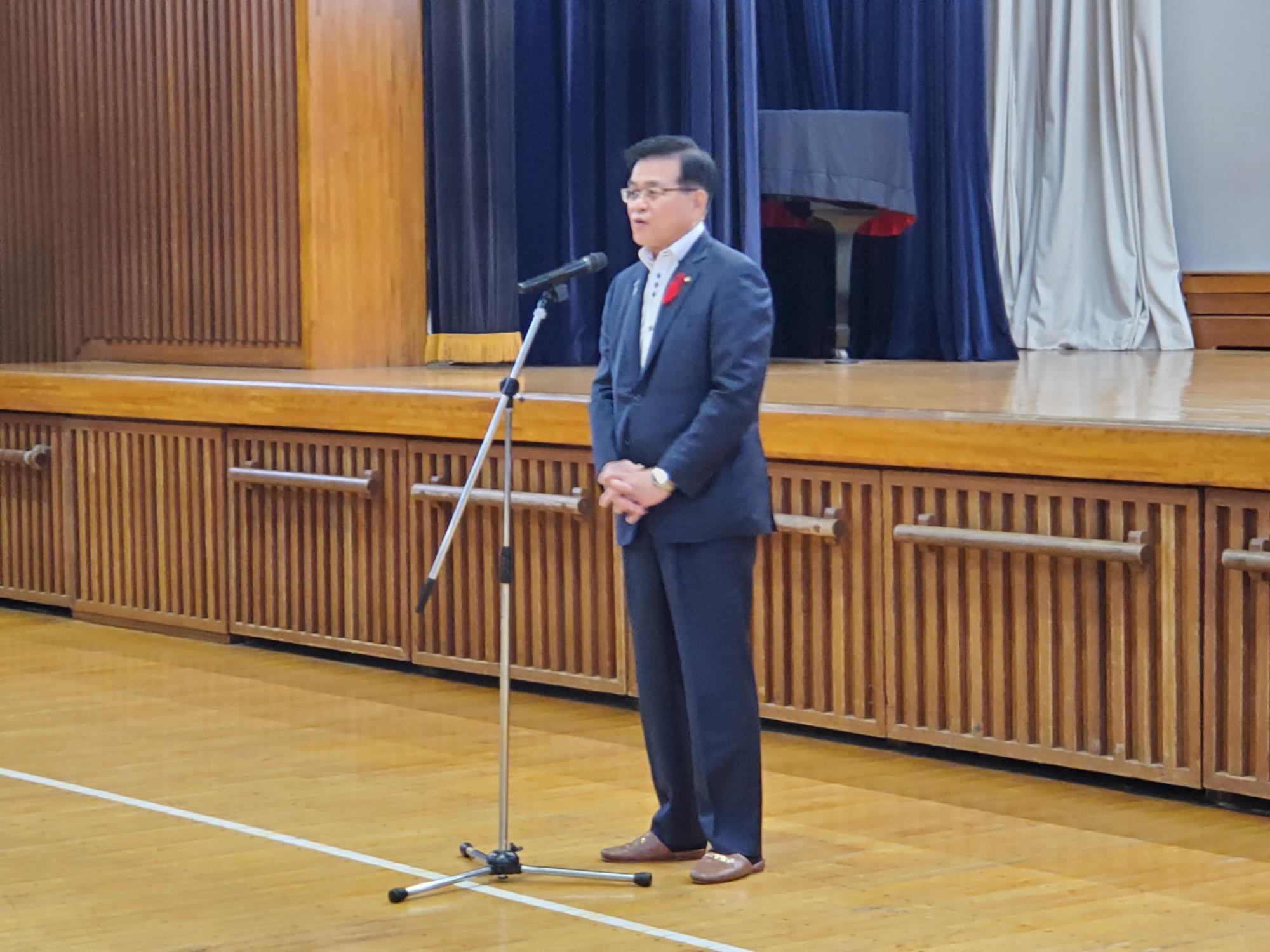 体育館のステージ前で、マイクスタンドで話をする高橋市長の写真